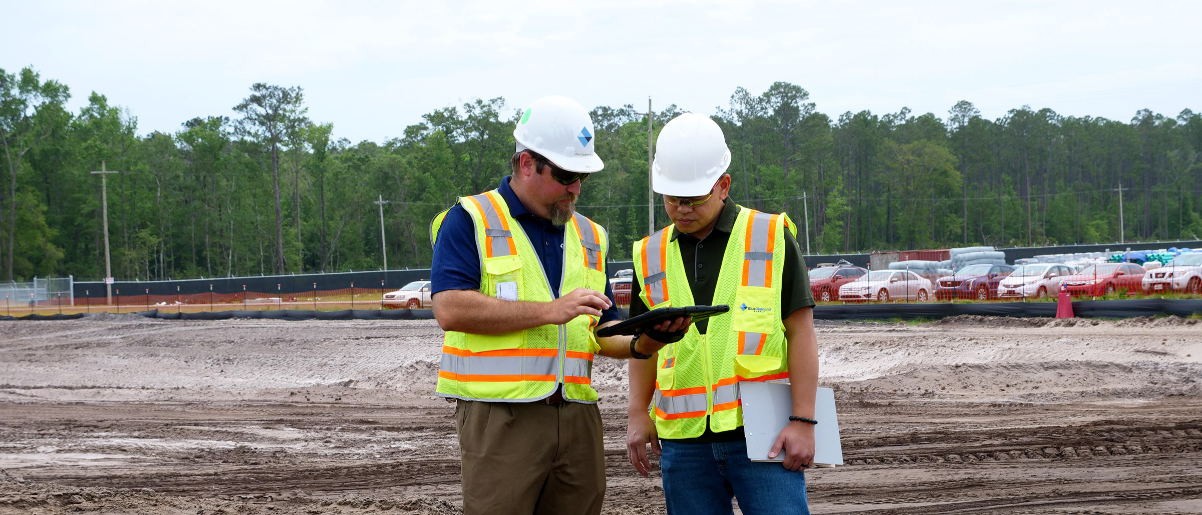 Two project team members in hard hats and safety vests reviewing plans on a tablet at a jobsite